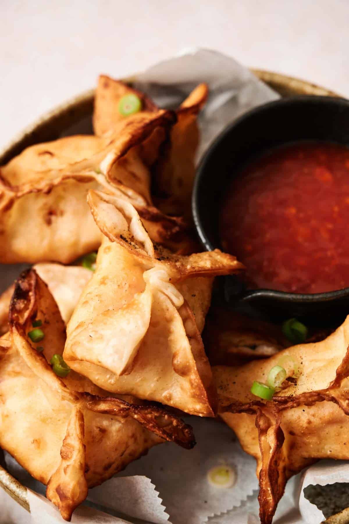 A plate of crispy crab rangoon fried wontons garnished with green onions, served with a small bowl of red dipping sauce.