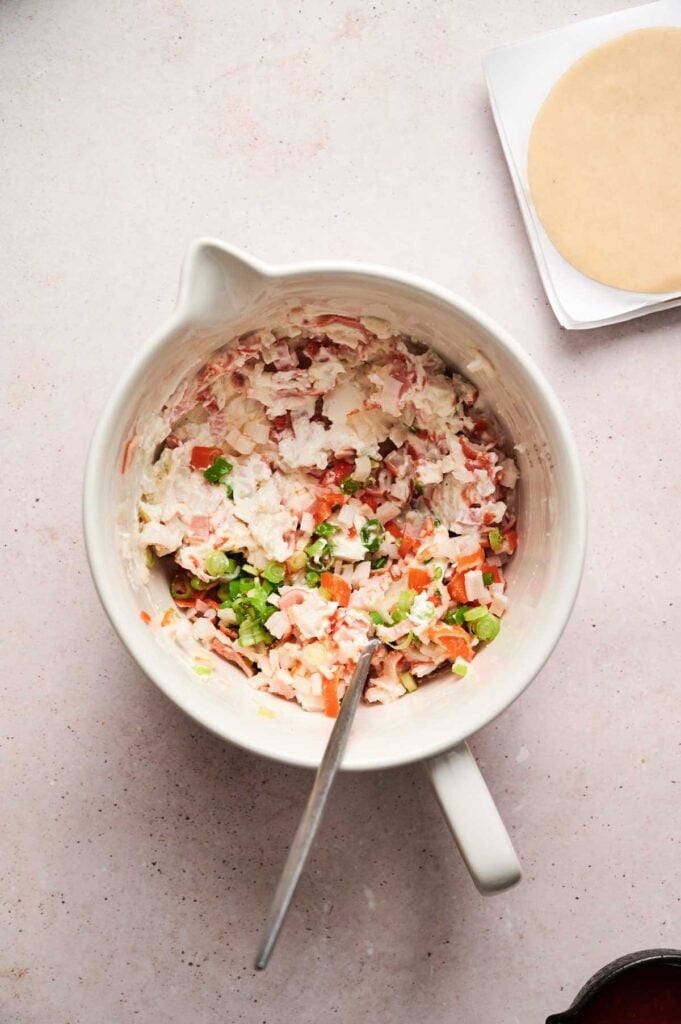 A bowl of creamy crab rangoon filling with chopped vegetables and green onions, with a spoon inside. A stack of round dough wrappers is visible in the corner.
