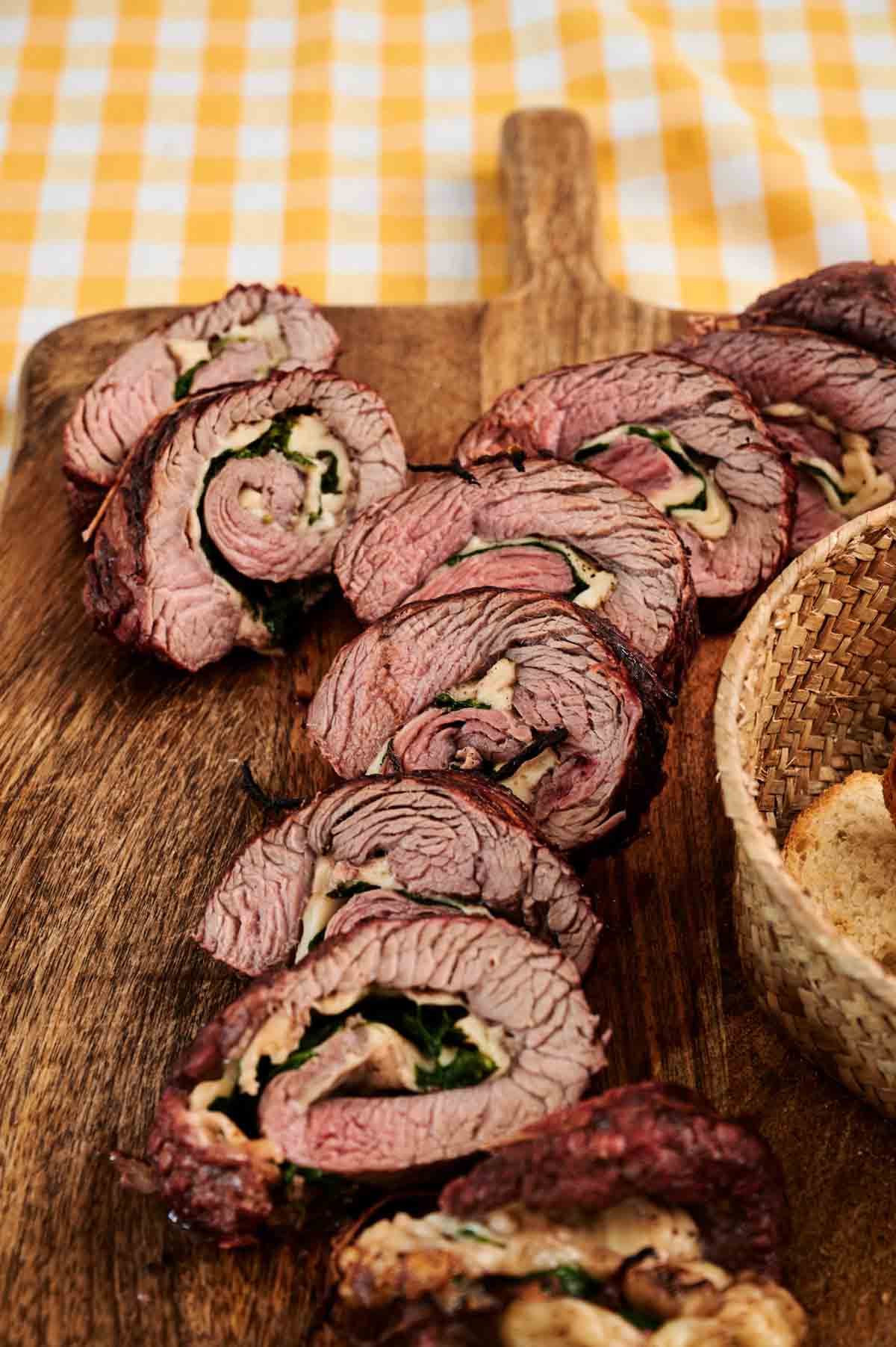 Grilled Steak Pinwheels stuffed with spinach and cheese are sliced and arranged on a wooden cutting board, with part of a woven basket visible on the side.