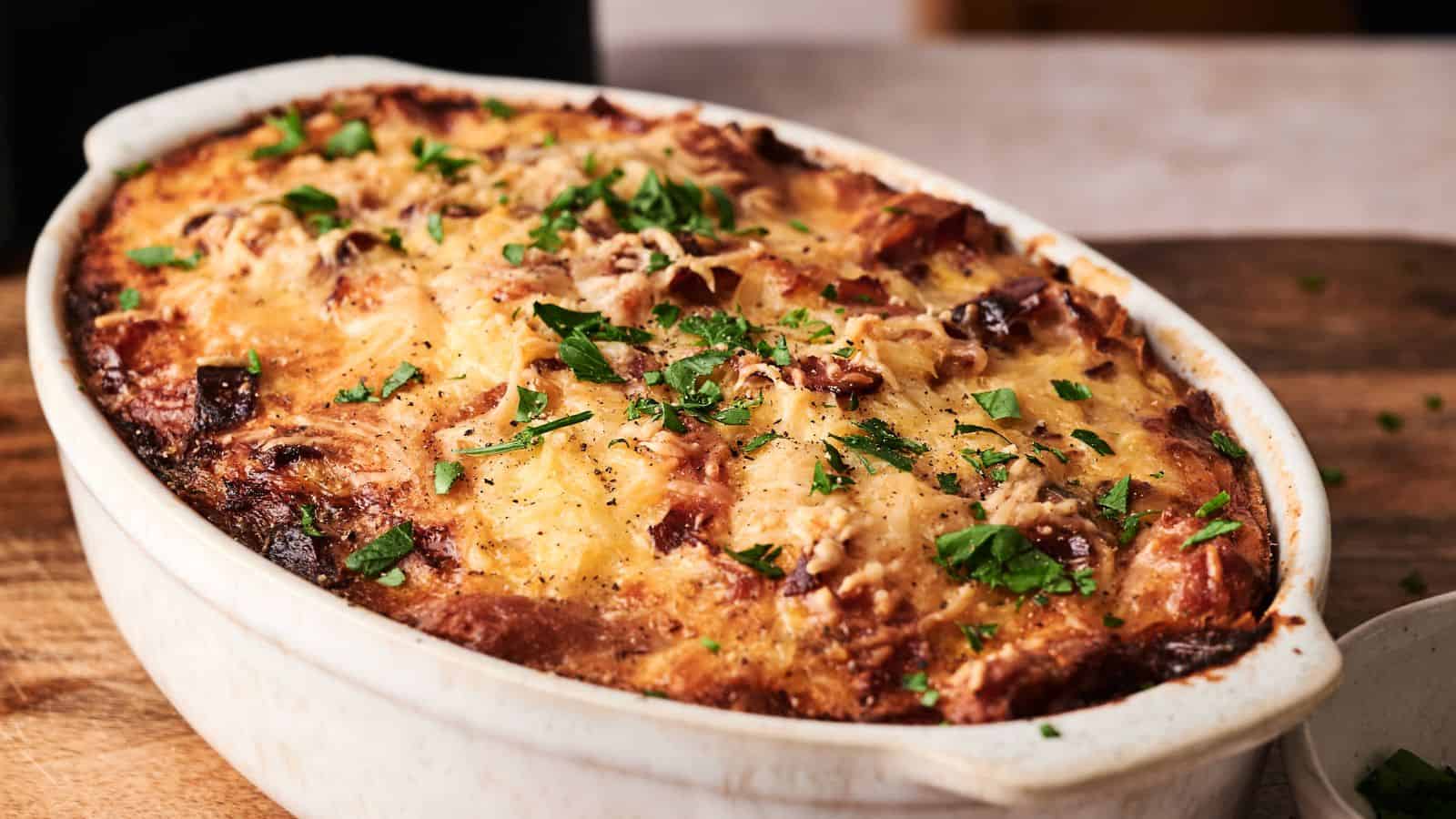 A baked casserole in an oval white dish, topped with melted cheese and chopped fresh herbs, sits on a wooden surface.