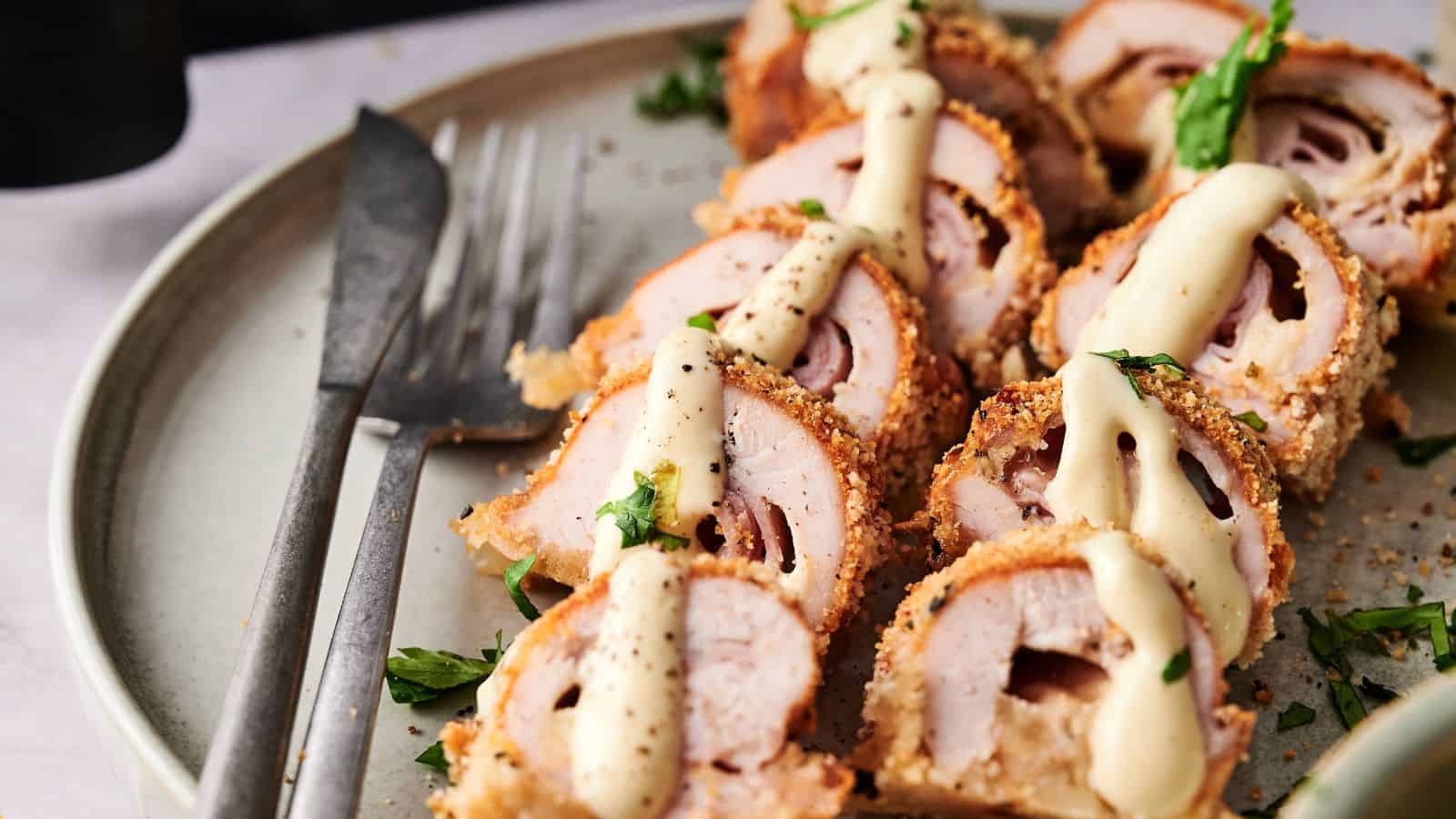 Sliced breaded chicken rolls topped with a creamy sauce and garnished with herbs, served on a plate with a fork and knife.