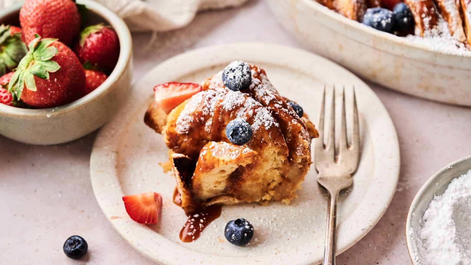 A serving of French toast casserole topped with blueberries, strawberries, and powdered sugar sits on a plate next to a fork; a bowl of fresh strawberries is nearby.