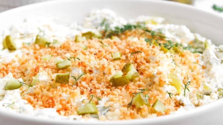 A close-up of a creamy dip topped with golden breadcrumbs, chopped pickles, and fresh dill in a white dish.