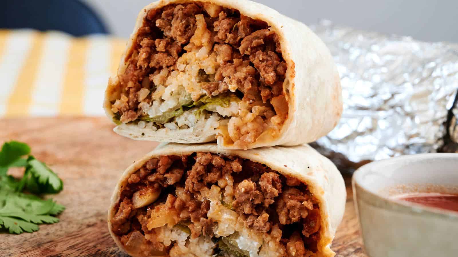 A burrito cut in half showing layers of seasoned ground beef, rice, cheese, and vegetables, with foil and a small bowl of sauce in the background.