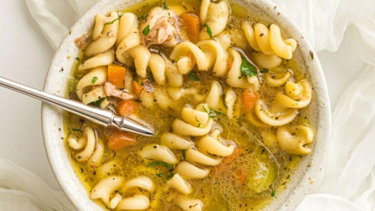 A bowl of rotini pasta soup with chunks of chicken, carrots, and herbs, with a spoon resting inside.