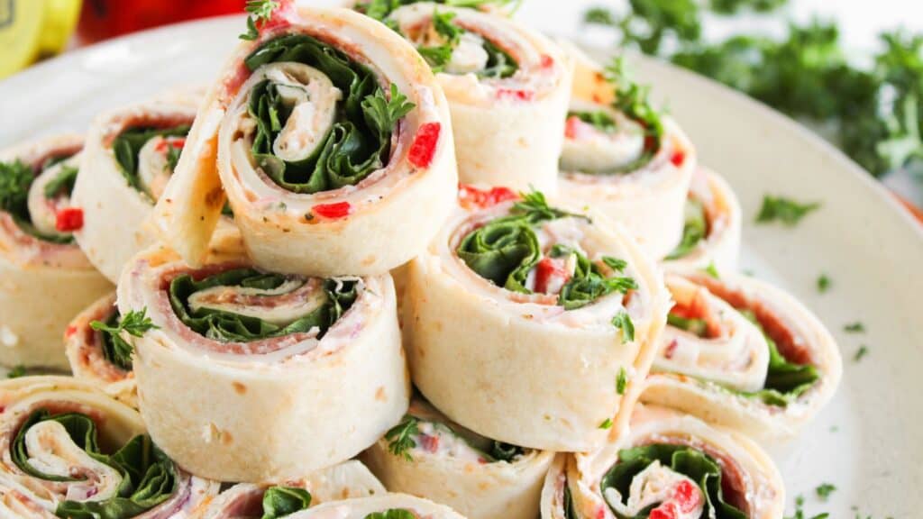 A plate of tortilla pinwheel sandwiches filled with spinach, diced red peppers, and cream cheese, garnished with fresh parsley.