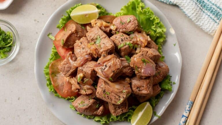 A plate of stir-fried beef cubes with onions, garnished with herbs, lettuce, tomato slices, and lime wedges, served on a white plate with chopsticks beside it.