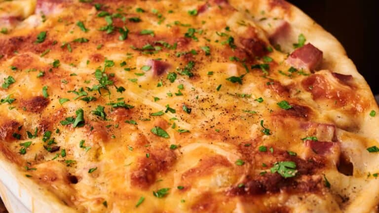 A close-up of a baked casserole topped with melted cheese, diced ham, visible seasoning, and chopped parsley.