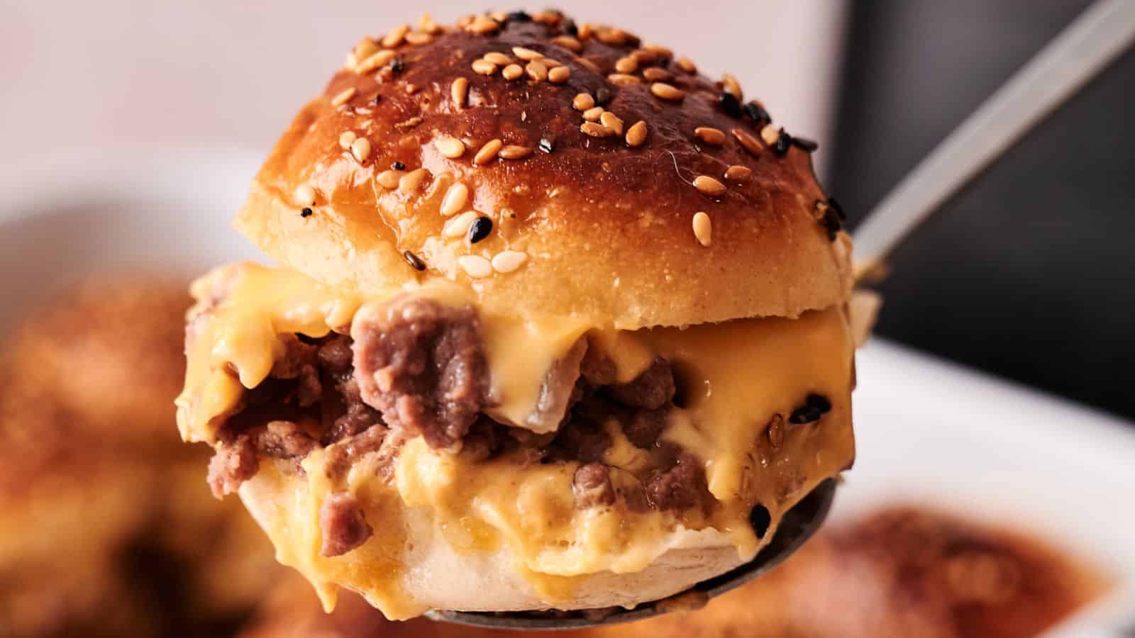 A close-up of a hamburger slider topped with sesame seeds, filled with ground beef and melted cheese, being lifted by a metal spatula.
