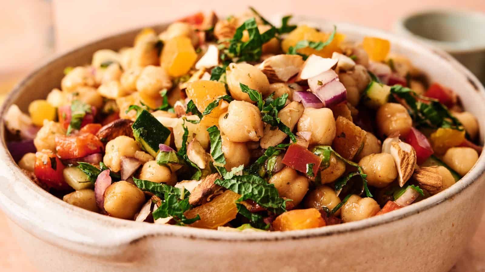 A ceramic bowl filled with chickpea salad, including chopped red onion, yellow peppers, tomatoes, almonds, and fresh herbs.