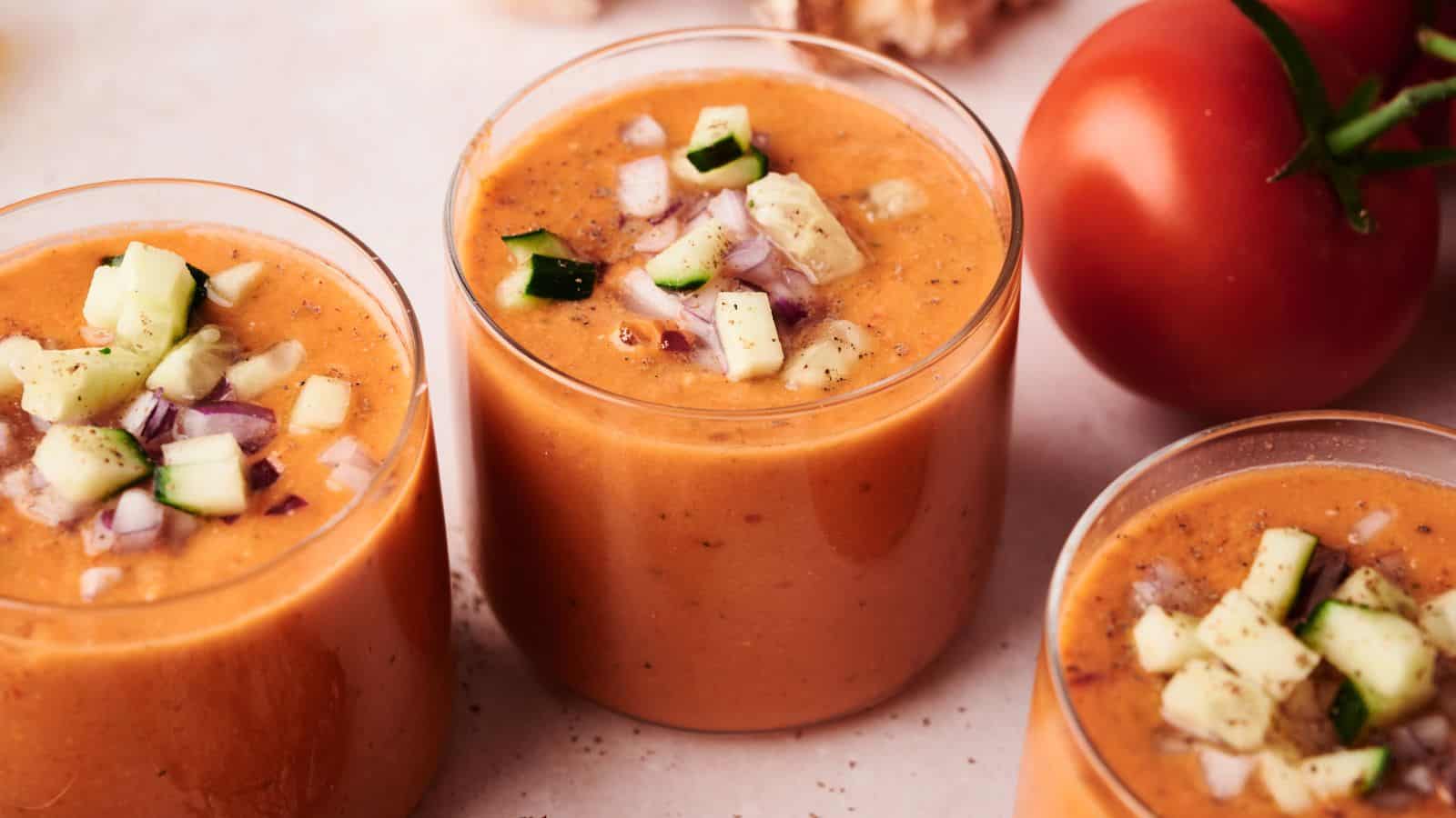 Three glasses of gazpacho soup topped with chopped cucumber and red onion, with a whole tomato in the background on a light surface.