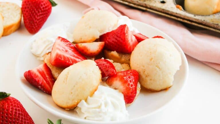 A plate of shortcake biscuits topped with sliced strawberries and dollops of whipped cream, with whole strawberries and a muffin tin in the background.