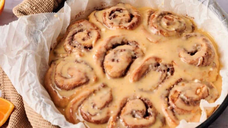 A round baking dish lined with parchment paper holds several cinnamon rolls topped with a generous layer of glaze.