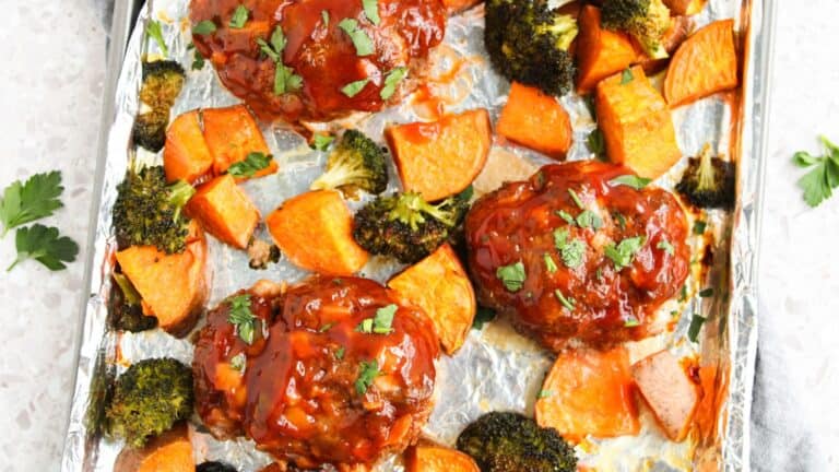 Three mini meatloaves topped with sauce are on a foil-lined baking sheet with roasted broccoli and sweet potato chunks, garnished with chopped herbs.