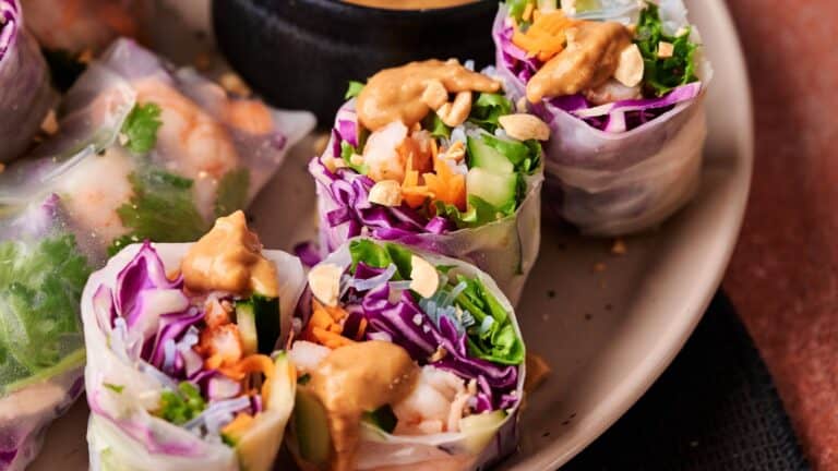 Fresh spring rolls filled with shrimp, lettuce, purple cabbage, carrots, and herbs are topped with peanut sauce and chopped peanuts, served on a plate.