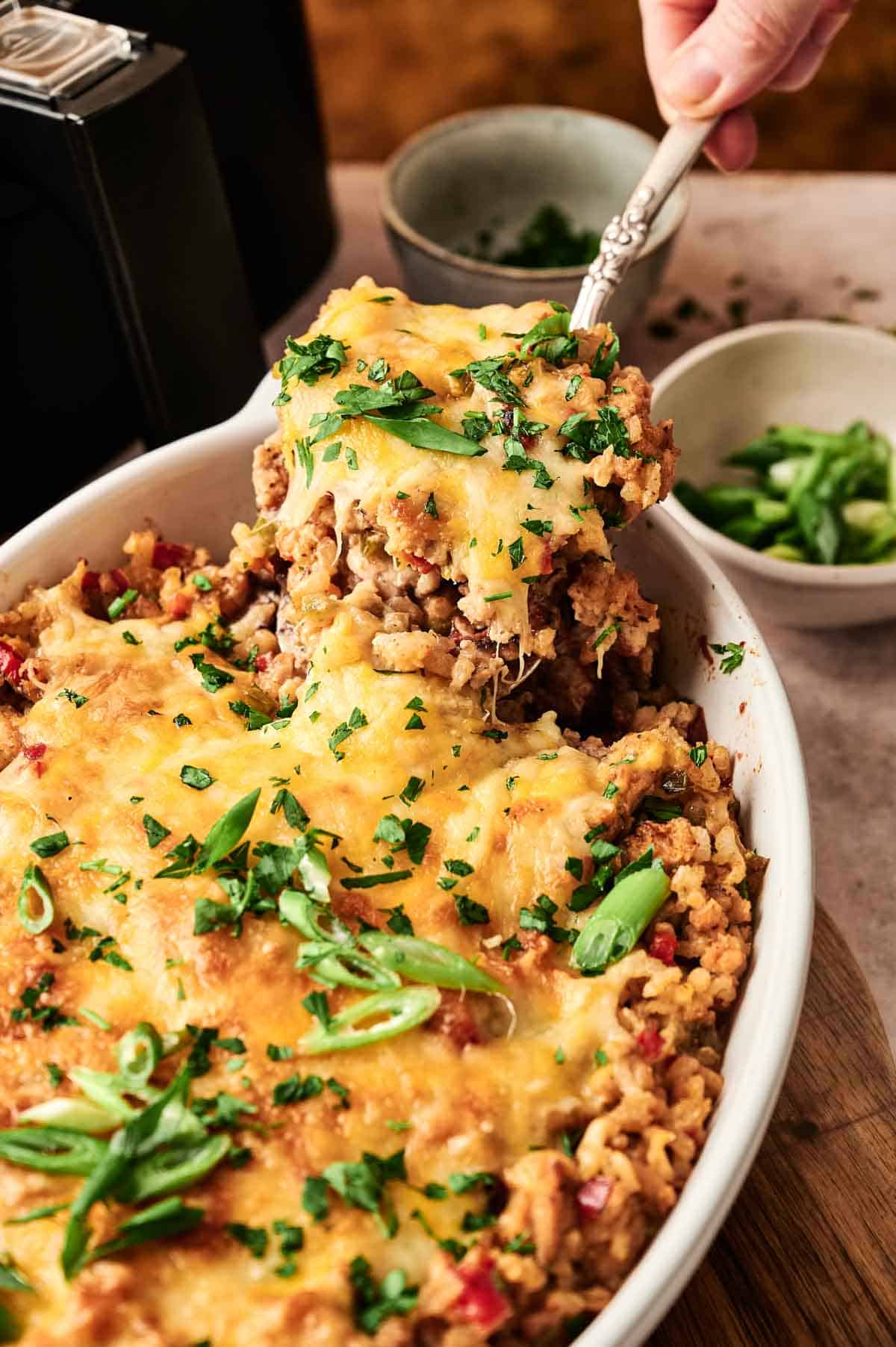 A hand lifts a serving of cheesy Air Fryer Mexican Casserole with ground meat, rice, and chopped green onions from a white dish.