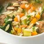 A bowl of Slow Cooker Italian Wedding Soup featuring meatballs, sliced almonds, chopped carrots, celery, spinach, and herbs in a clear broth.