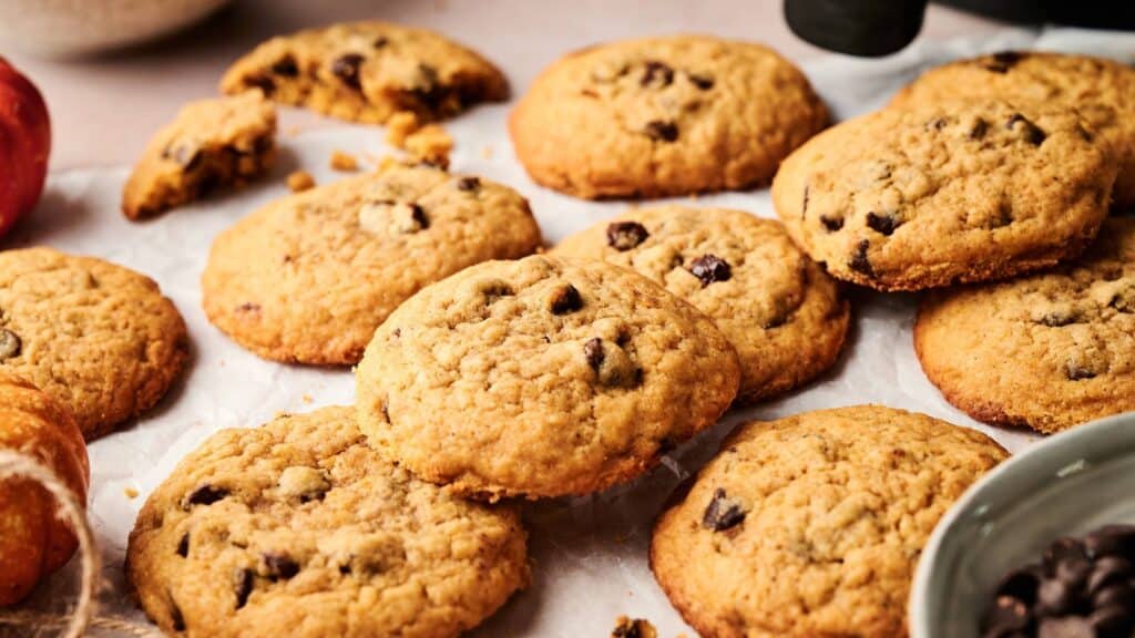 A batch of chocolate chip cookies is arranged on parchment paper, with some whole and some partially broken, surrounded by bowls and other kitchen items.