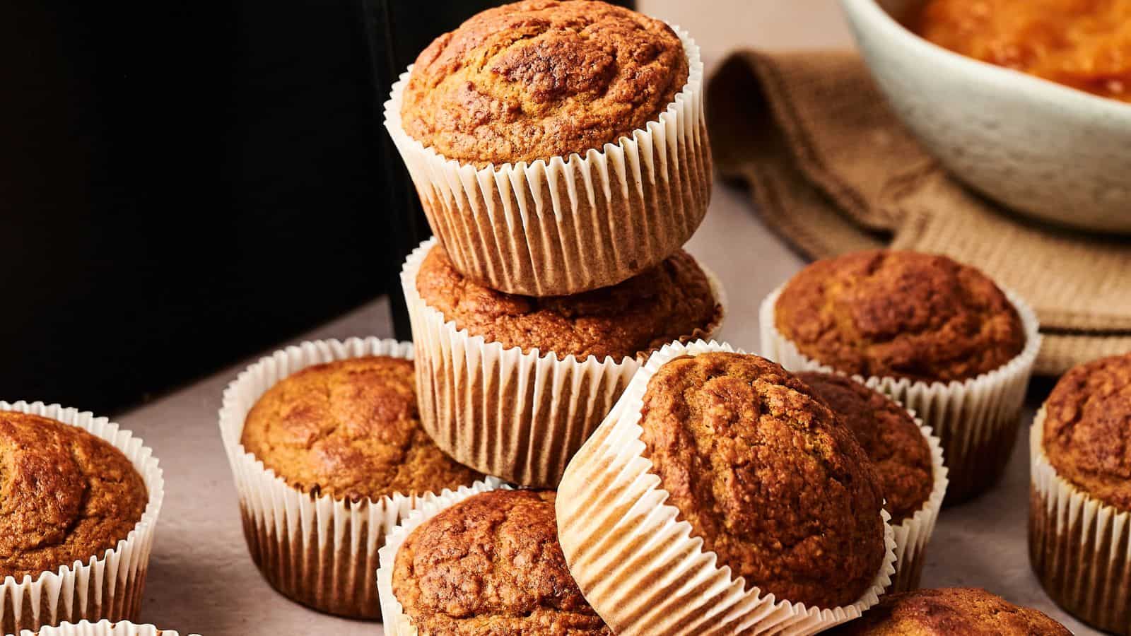 A stack of golden-brown muffins in white paper liners is arranged on a table next to a bowl and a brown cloth.