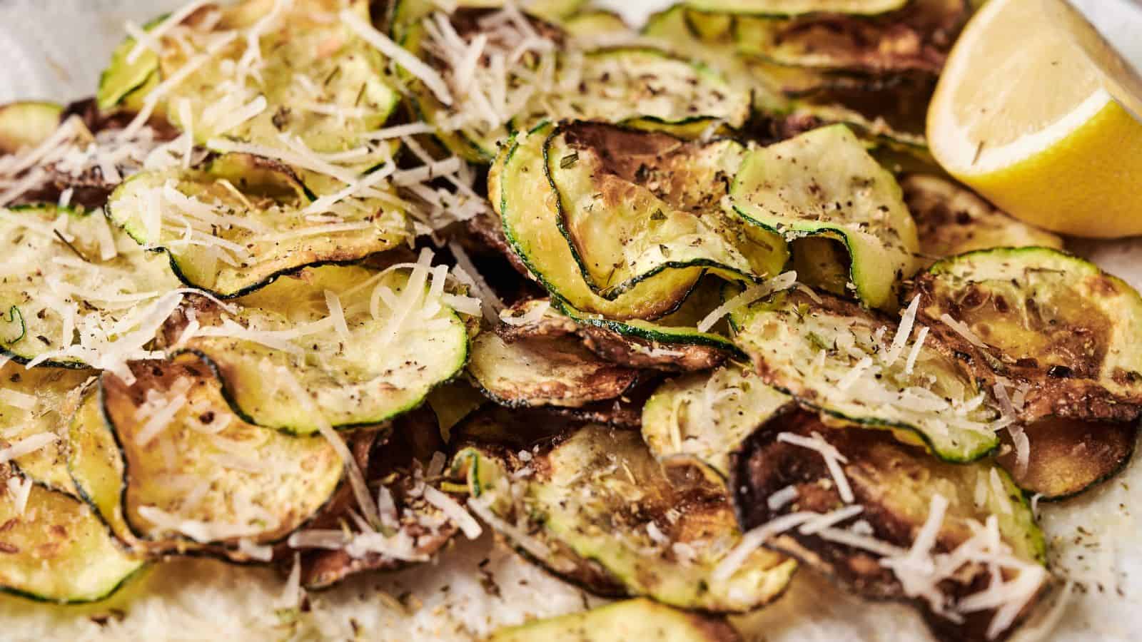 A plate of baked zucchini chips topped with shredded parmesan cheese and a lemon wedge on the side.
