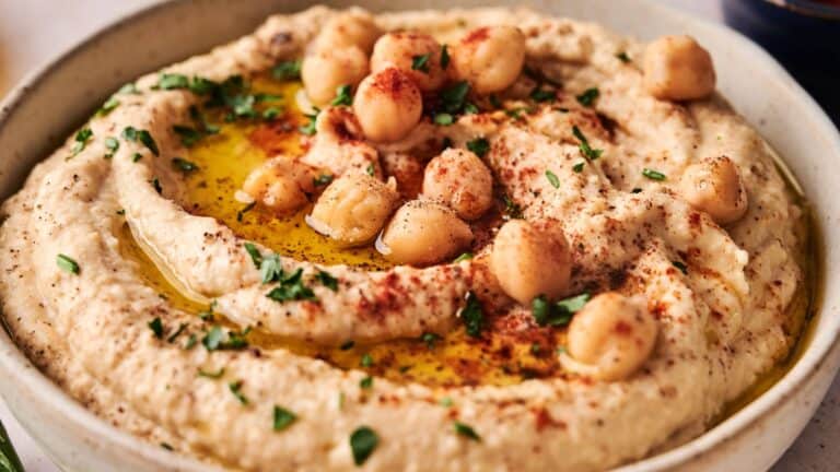 A bowl of hummus topped with whole chickpeas, chopped parsley, olive oil, and a sprinkle of paprika.