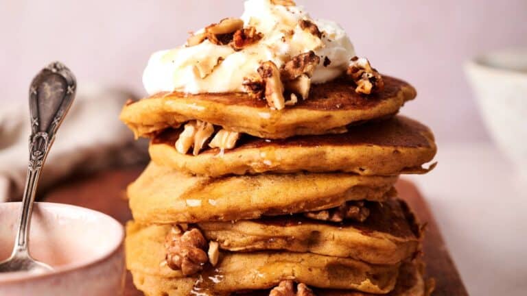 A stack of pancakes topped with whipped cream, chopped walnuts, and syrup, served on a wooden board with a spoon and bowl nearby.