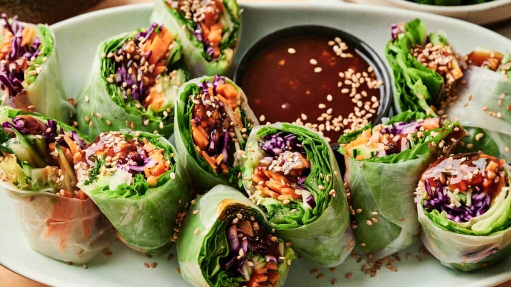 A plate of fresh vegetable spring rolls filled with greens, shredded carrots, and purple cabbage, served with a bowl of dipping sauce and sprinkled with sesame seeds.