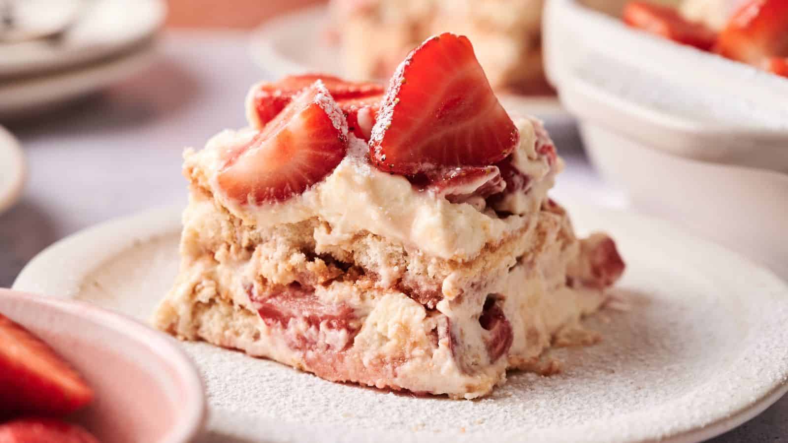 A slice of strawberry tiramisu with layers of cream, ladyfingers, and fresh strawberries is served on a white plate.