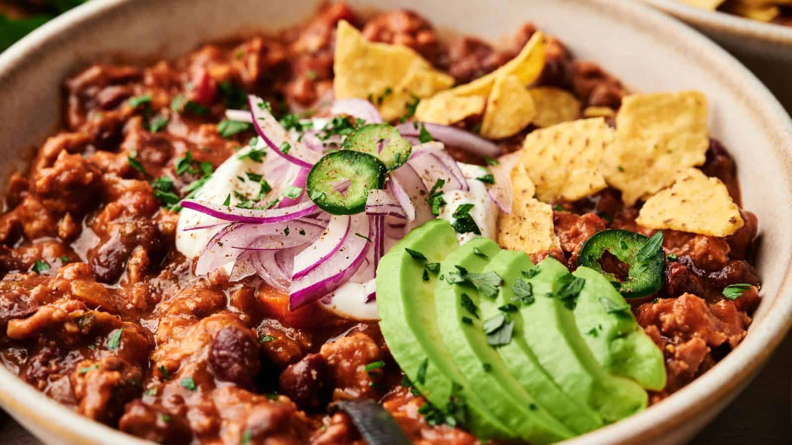 A bowl of chili topped with sliced avocado, red onions, jalape&ntilde;os, sour cream, chopped herbs, and tortilla chips.