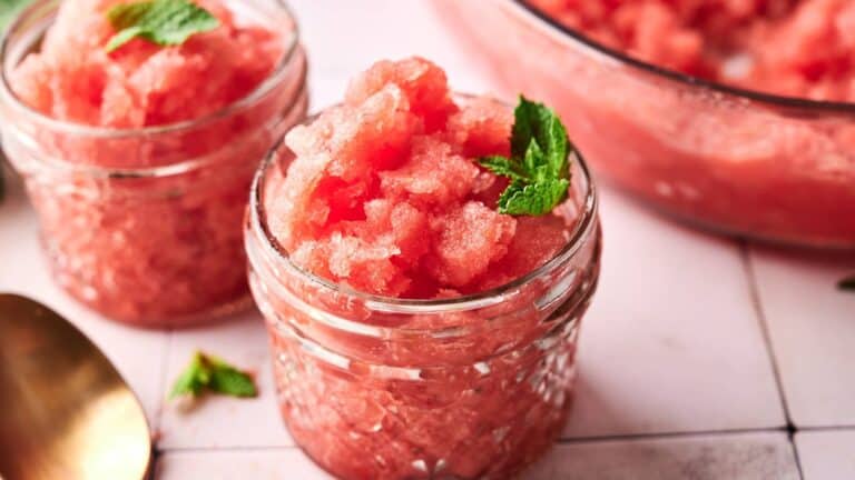 Two small glass jars filled with pink watermelon granita, garnished with fresh mint leaves, sit on a white surface next to a gold spoon.