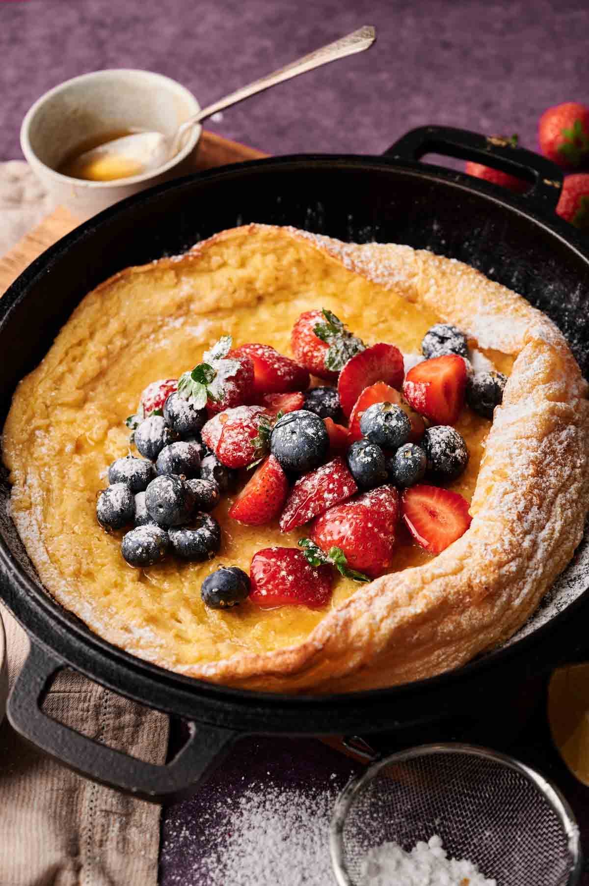 A German Pancake, or Dutch baby, in a cast iron skillet topped with strawberries, blueberries, powdered sugar, and mint, with honey and a sieve nearby.