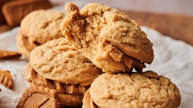 A stack of sandwich cookies with a light brown filling sits on parchment paper; one cookie has a bite taken out of it.