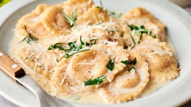 A plate of ravioli in a creamy sauce, topped with grated cheese and fresh herbs, with a fork resting on the side.