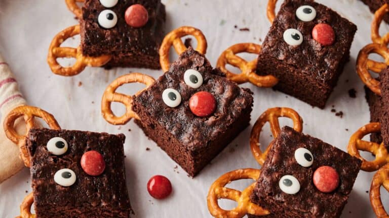 Brownie squares decorated with candy eyes, red candies for noses, and pretzels for antlers, arranged on a white surface to resemble reindeer faces.