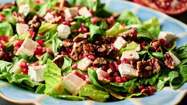 A salad with chopped romaine lettuce, feta cheese cubes, pecans, and pomegranate seeds on a blue plate.