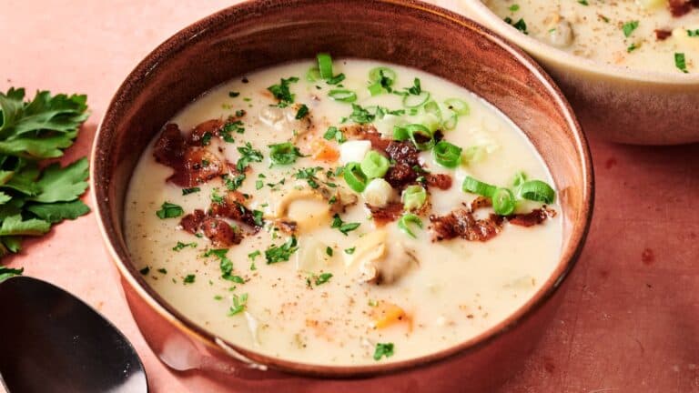 A bowl of creamy soup with chopped green onions, herbs, bacon pieces, and vegetables, placed on a pink surface next to a spoon and fresh parsley.