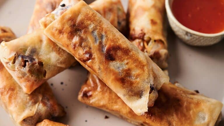 Several golden-brown egg rolls are stacked on a plate next to a small bowl of red dipping sauce.