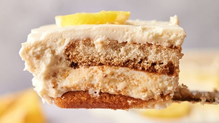 A close-up of a slice of layered tiramisu topped with a lemon wedge, showing creamy and cake-like textures on a fork.
