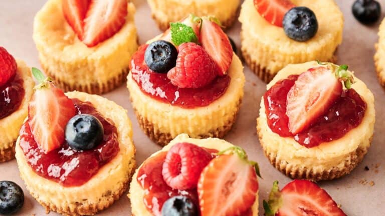 Mini cheesecakes topped with strawberry sauce, fresh strawberries, blueberries, raspberries, and mint leaves are arranged on a light surface.