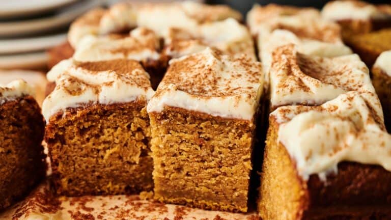 Close-up of sliced pumpkin cake bars topped with cream cheese frosting and a dusting of cinnamon, arranged on a parchment-lined surface. Plates are visible in the background.