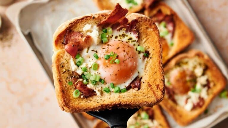A slice of toasted bread with a cooked egg, bacon, white cheese, and chopped green onions on top, held on a spatula above a tray with similar slices.
