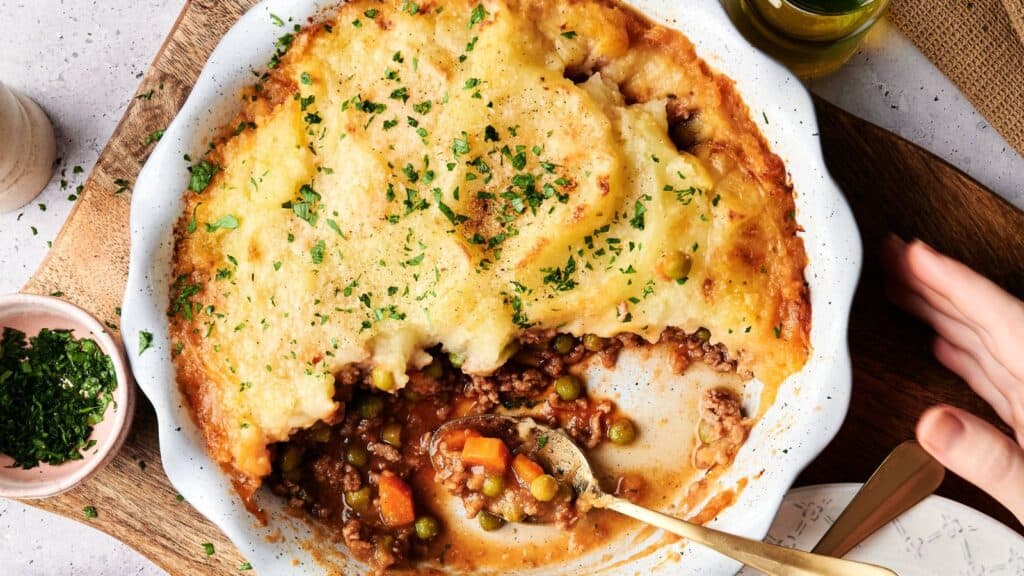 A partially served shepherd&rsquo;s pie in a white dish, showing mashed potato topping and a filling of ground meat, peas, carrots, and sauce. A spoon rests in the dish.