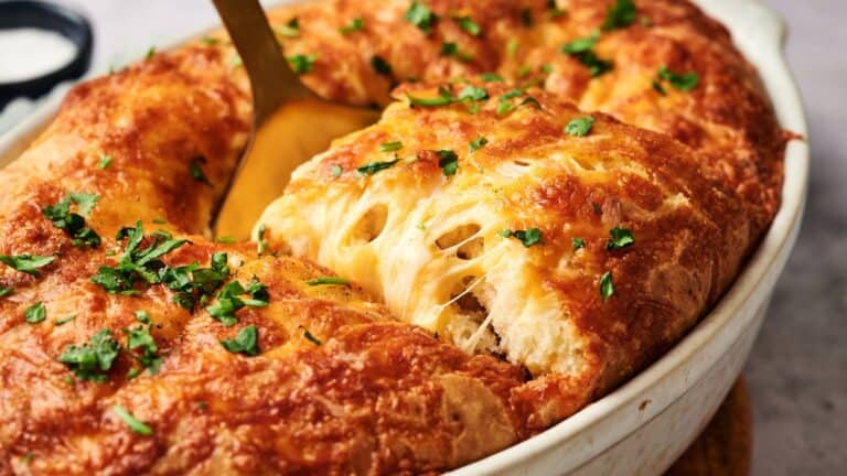 A close-up of a cheesy baked bread dish in a white oval baking dish, with a serving spoon lifting a piece and chopped herbs sprinkled on top.
