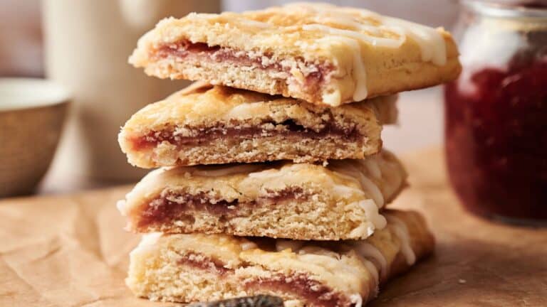 Four rectangular pastries with a fruit filling and white icing are stacked on brown parchment paper, with a jar of jam in the background.