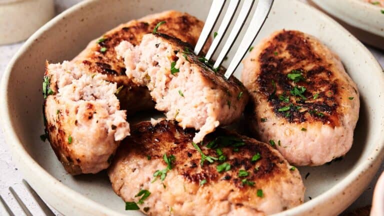 Four browned sausage patties garnished with chopped herbs, with one patty cut open by a fork to show the cooked interior, served in a beige bowl.