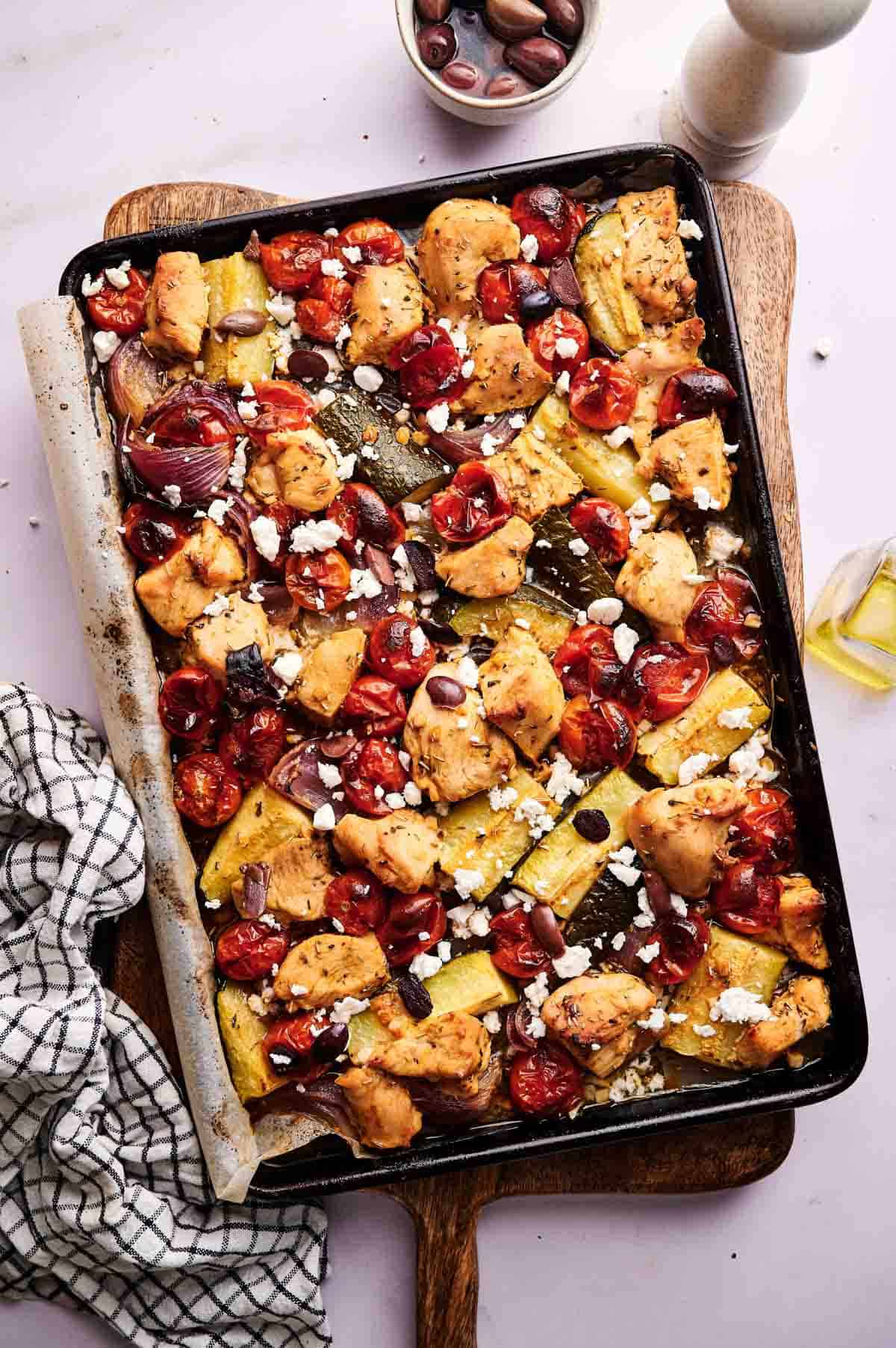 A Greek Sheet Pan Chicken dinner with baked chicken pieces, cherry tomatoes, zucchini, red onions, olives, and crumbled feta cheese sits on a wooden board next to a checked towel.