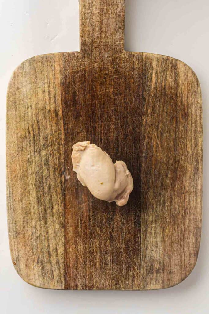 A single piece of cooked chicken breast, perfect for shredding into a hearty Slow Cooker White Chicken Chili, rests in the center of a worn wooden cutting board.
