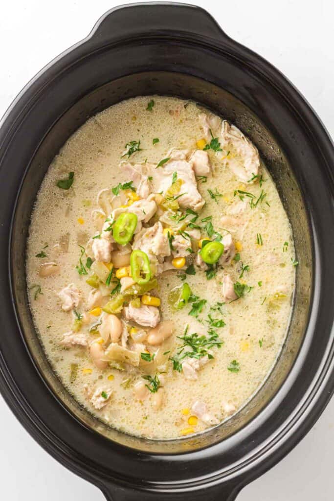 A slow cooker filled with Slow Cooker White Chicken Chili, featuring tender chunks of chicken, white beans, corn, chopped herbs, and sliced green peppers for a creamy and comforting meal.