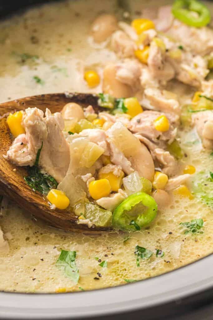 A wooden spoon lifts a portion of Slow Cooker White Chicken Chili, brimming with creamy chicken, corn, white beans, onions, celery, and jalape&ntilde;o slices from a pot.