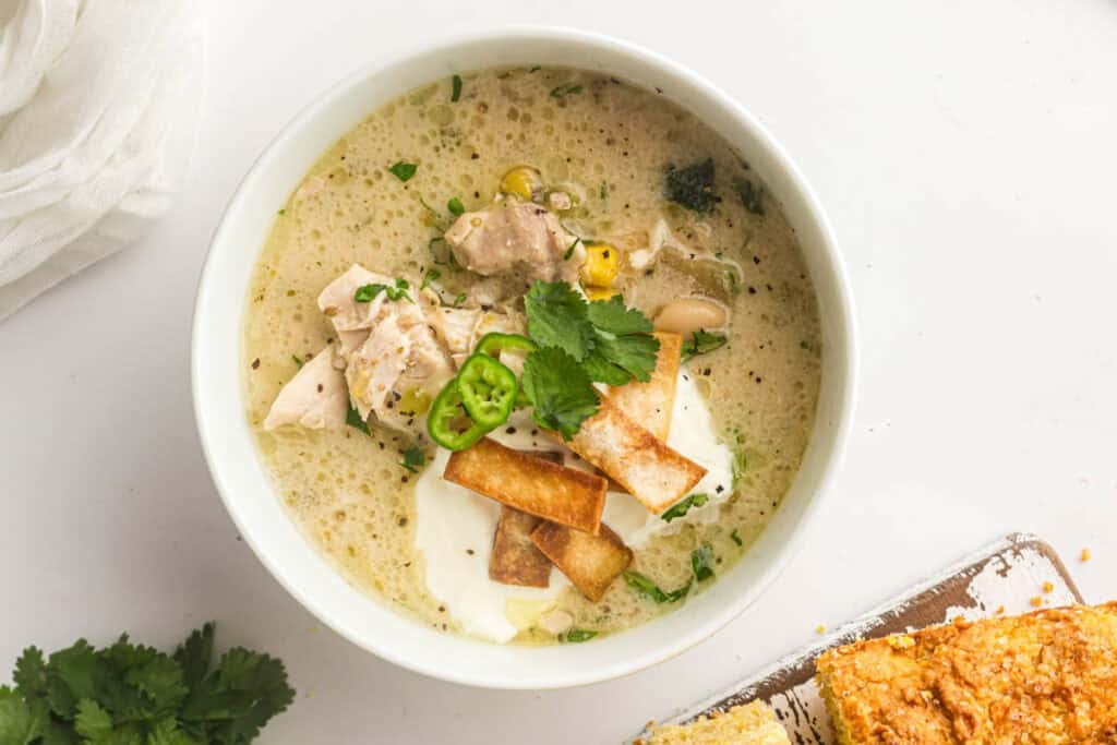 A bowl of Slow Cooker White Chicken Chili topped with tortilla strips, sliced jalape&ntilde;o, cilantro, and sour cream sits beside a piece of cornbread.